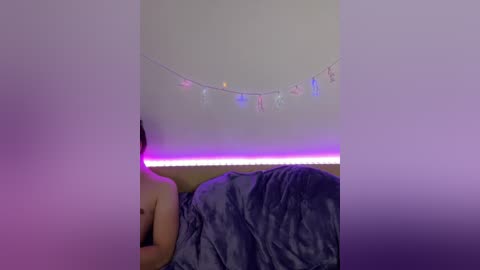 Video of a nude, light-skinned person with dark hair lying on a bed, partially covered by a dark gray blanket. The room has a purple and white color scheme with a string of colorful lights on the ceiling.