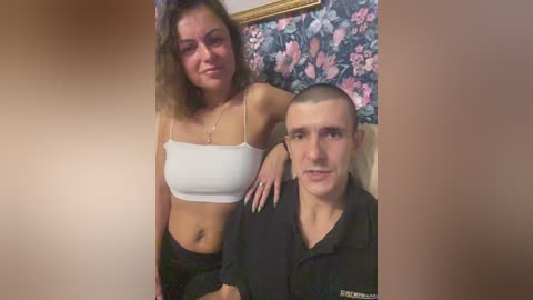 Video of a young woman with curly brown hair in a white crop top, standing next to a bald man in a black hoodie, both smiling, in a room with floral wallpaper.