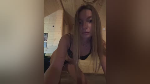 Video of a young, slim, fair-skinned woman with long, straight blonde hair, wearing a black bra, leaning forward in a wooden cabin.
