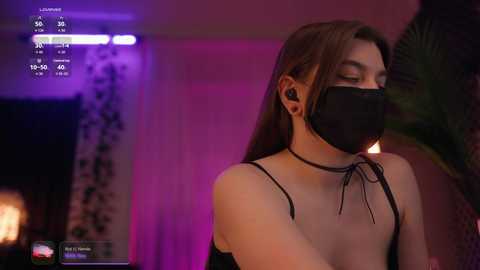 Video of a slender, light-skinned woman with straight brown hair, wearing a black face mask, black choker, and black tank top, standing in a dimly lit room with purple lighting and a palm tree in the background.