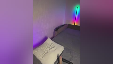 Video of a dimly lit bedroom with a gray bed, a beige pillow, and a tall, colorful LED lamp emitting green, blue, and red hues on the right wall.
