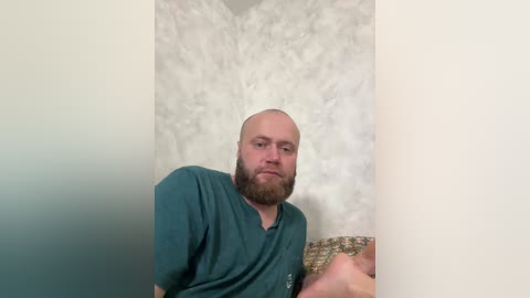 A video of a bald, bearded man with a contemplative expression, wearing a green shirt, standing in front of a textured, white wall.