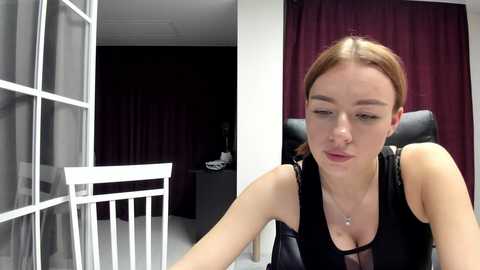 Video of a pale-skinned, light-haired woman in a black tank top, seated in a chair, with a white baby crib and dark curtains in the background.