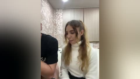 A video of a young woman with long, blonde hair in pigtails, wearing a white turtleneck sweater, sitting in a room with patterned wallpaper and vertical blinds.