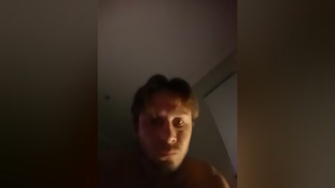 A blurry, low-light video of a young man with light skin and short hair, wearing a dark shirt, standing in a dimly lit room with a dark wall and shadowy corners.