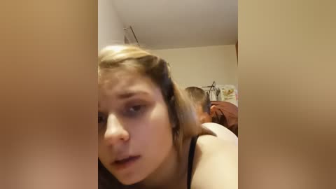 A blurry video of a young woman with light skin and shoulder-length blonde hair, wearing a black tank top, sitting on a bed in a dimly lit room.