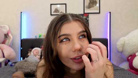 Video of a young woman with fair skin, brown hair, and large blue eyes, lying on a bed with plush toys, framed art, and soft lighting in the background.