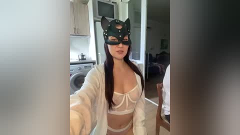 Video of a woman in white lingerie and a black cat mask, standing in a modern kitchen. She has long brown hair and fair skin, and her breasts are visible through the sheer fabric.