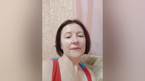 Video of a middle-aged, Caucasian woman with fair skin and shoulder-length brown hair, wearing a red tank top, smiling in a room with beige textured wallpaper and pink curtains.