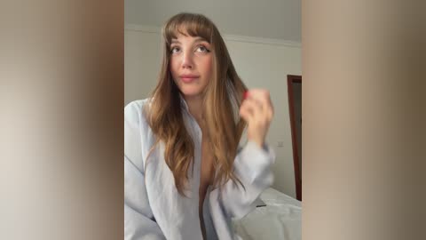A candid video of a Caucasian woman with long, wavy brown hair, wearing a light-colored robe, standing in a simple bedroom. She looks surprised and playful, holding a strand of her hair.