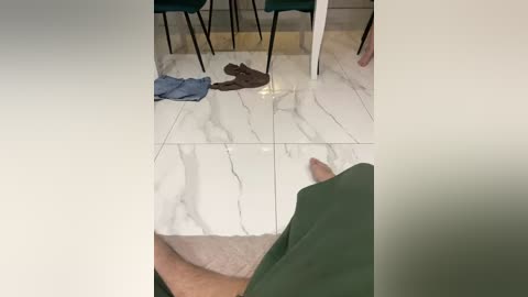 A video captures a man's legs and feet on a white marble floor with grey veining, next to scattered clothes including blue jeans and brown shorts. The background shows a modern, minimalistic room with green chairs and a white table.