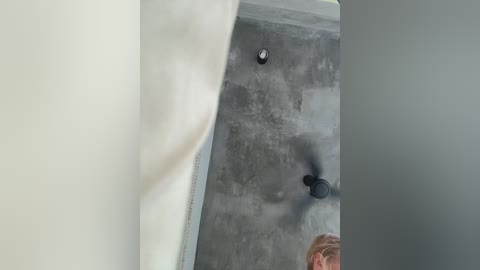 A video of a person in a black jacket and blonde hair walking on a concrete floor with a textured wall and light fixtures in a modern indoor setting.