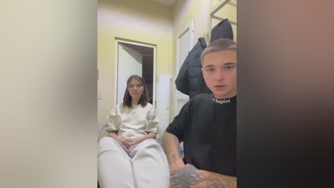 Video of two people in a hallway: a woman with shoulder-length brown hair in white scrubs, and a tattooed man in a black shirt.
