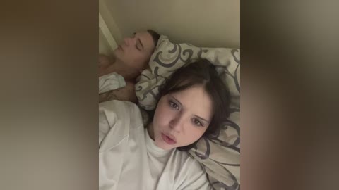 Video of a man and woman in bed, both with fair skin. Woman with long brown hair, wearing a white nightgown, gazes at man, who is shirtless with short brown hair, lying next to her.