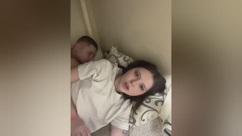 A video of a young woman with shoulder-length dark hair, wearing a beige shirt, lying on a bed with a patterned pillow, while a young boy sleeps next to her. The room is dimly lit, and the scene is intimate.