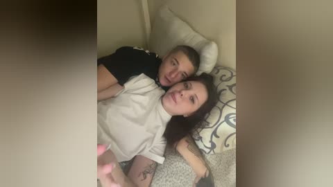 A video of a young Caucasian couple lying on a bed with beige walls. The man, with short blond hair, wears a black shirt; the woman, with long dark hair, is in a white turtleneck.