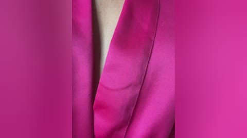 A close-up video of a woman's neck and chest, covered in vibrant magenta satin fabric, creating a smooth, glossy texture. The background is blurred, focusing solely on the fabric's rich color and soft folds.