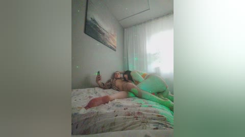 Video of two nude women, one lying on a bed, the other sitting beside her, holding a phone, in a dimly lit room with a green glow and a painted seascape on the wall.
