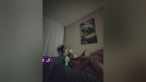 A dimly lit video captures a woman lying on a gray couch, wearing a white shirt and beige hat, with a tattooed arm. A large, abstract eye painting hangs on the beige wall.