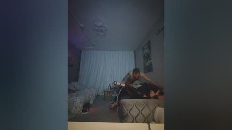 Video of a dimly lit bedroom with white curtains, two beds, a man in a tank top and jeans, and a woman in a dress.