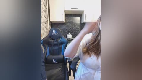 A video of a woman in a white shirt standing in a modern kitchen with a black granite countertop, white cabinets, and a brick wall. She is holding a smartphone near her face.