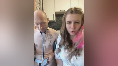 Video of a bald man wearing a stethoscope, with a young woman wearing a white shirt and pink feather boa, standing in a modern kitchen with white cabinets and a dark countertop.