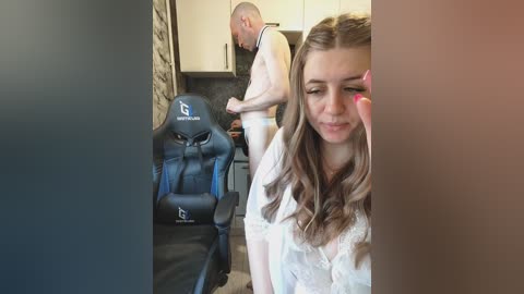 Video of a young woman in a white shirt, using a pink vibrator, with a shirtless man in the background in a modern kitchen.