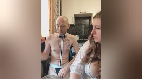 A video of a topless, bald man in a white doctor's coat and stethoscope, sitting on a chair beside a woman in a white lace bra, in a modern kitchen with white cabinets and a dark granite countertop.