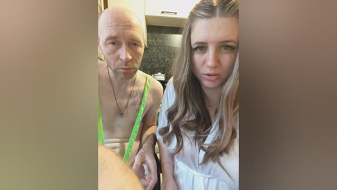 A video features a bald, shirtless man in green suspenders and a woman with long, wavy hair in a white blouse. They sit in a modern kitchen with a dark backsplash.