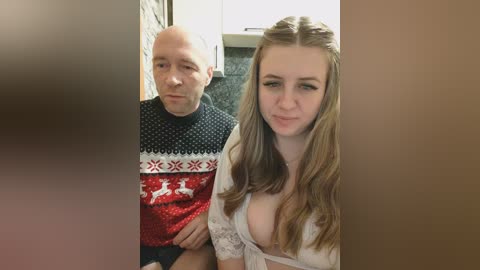 Video of a man and woman, both Caucasian, in a bathroom. The man, bald, wears a red sweater with a reindeer design. The woman, with long brown hair, wears a revealing white lace top, exposing cleavage.