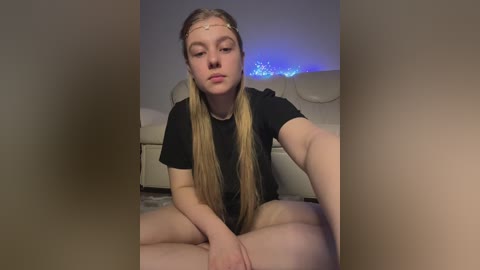 Video of a young, fair-skinned, blonde-haired woman in a black t-shirt, sitting cross-legged on a beige carpet, with a white couch and blue LED lights in the background.