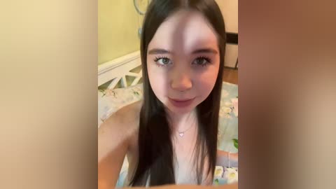 Video of a young Asian woman with long black hair and light skin, wearing a white top, sitting on a bed with a floral comforter.