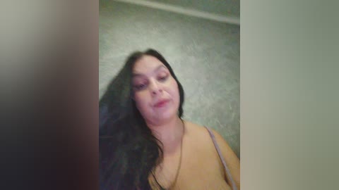 Video of a young woman with long black hair and fair skin, wearing a light-colored spaghetti strap top, standing in a dimly lit room with a textured gray wall.