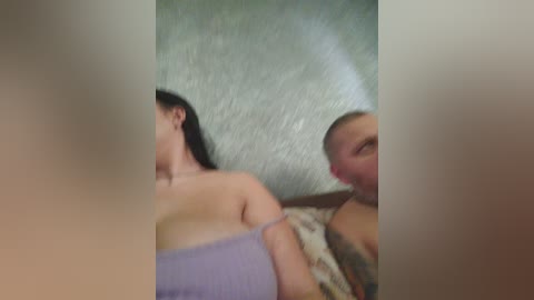 A blurry video of a man with a bald head and a woman with long black hair, both partially nude, lying on a bed with a floral-patterned blanket.