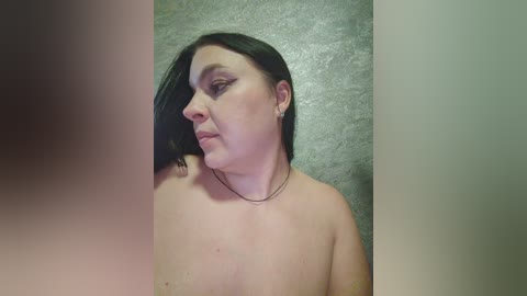 A video of a pale-skinned, middle-aged woman with shoulder-length dark hair, wearing minimal makeup and a thin necklace, standing against a textured, grayish wall. The image is cropped, with a blurred figure on the left.