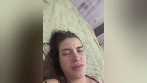 Video of a young woman with fair skin, long brown hair, and closed eyes lying on a green fuzzy blanket in a dimly lit room.