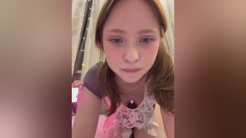 Video of a young woman with light skin, brown hair, and a slender physique, wearing a white lace top and necklace. She's indoors, with blurred background elements.