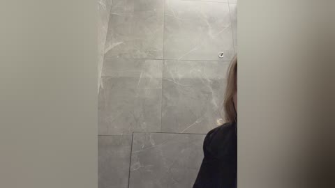 A video of a woman with long, straight hair, wearing a dark jacket, standing in a modern, minimalist bathroom with large, light gray marble tiles and a visible vent.