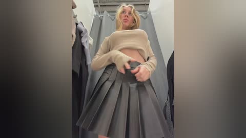 A low-angle video of a blonde woman in a beige sweater and grey pleated skirt, adjusting her skirt in a dressing room with grey curtains and white walls.