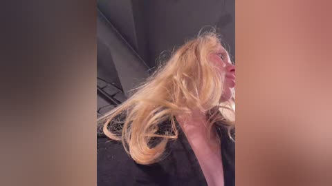 Video of a blonde woman with messy hair, wearing a black top, leaning against a dark background.