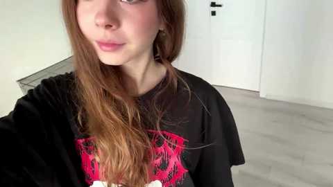 Video of a young Caucasian woman with long, wavy auburn hair, wearing a black graphic tee with pink details, in a modern, minimalistic room with light wooden flooring and white walls.