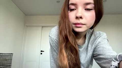 Video of a young Asian woman with long, straight brown hair, wearing a gray sweatshirt, looking contemplative, standing in a minimalist room with white walls and a closed door.