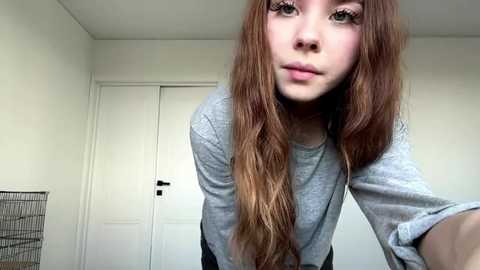 A video of a young woman with long, wavy auburn hair and light skin, wearing a gray long-sleeve shirt, standing in a sparsely decorated room with white walls and a closed door.