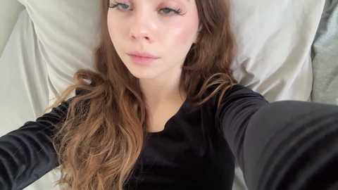 Video of a young woman with long, wavy brown hair, fair skin, and light makeup, lying on a white pillow, wearing a black velvet top, taking a selfie.