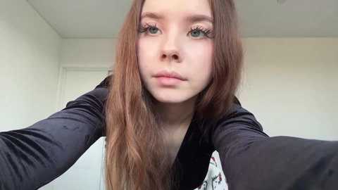 A video of a young Caucasian woman with long, straight brown hair, wearing a black velvet top, green eyes, and fair skin, in a plain white room with a closed door.