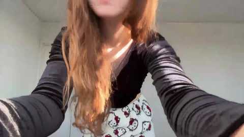 Video of a young woman with long, wavy, light brown hair, wearing a black long-sleeve top and a white skirt with a pattern of Hello Kitty faces. The background is a plain, white-walled room with minimal decor.