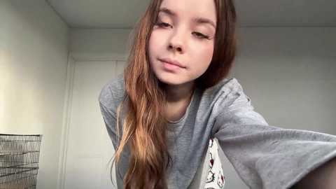 Video of a young, fair-skinned girl with long, wavy auburn hair, wearing a gray T-shirt, leaning slightly forward, indoors with white walls and a closed door in the background.