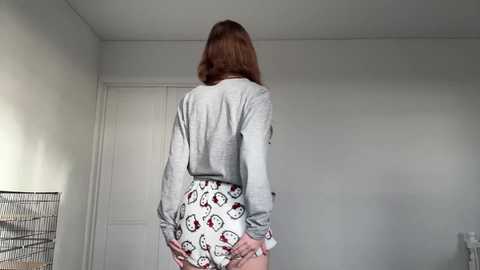 Video of a slender, light-skinned woman with shoulder-length auburn hair, wearing a gray, long-sleeve top and white panties with red and white cartoon characters, standing in a minimalist, white-walled room with a door and a wire mesh screen.