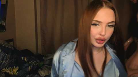 Video of a young woman with fair skin and long, straight brown hair, wearing a light blue shirt, sitting on a bed with a patterned blanket. The background features a dimly lit room with a curtain.