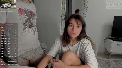 Video of a young woman in a white T-shirt and shorts sitting on a bed, looking serious, with a digital overlay of health stats, in a modern bedroom.